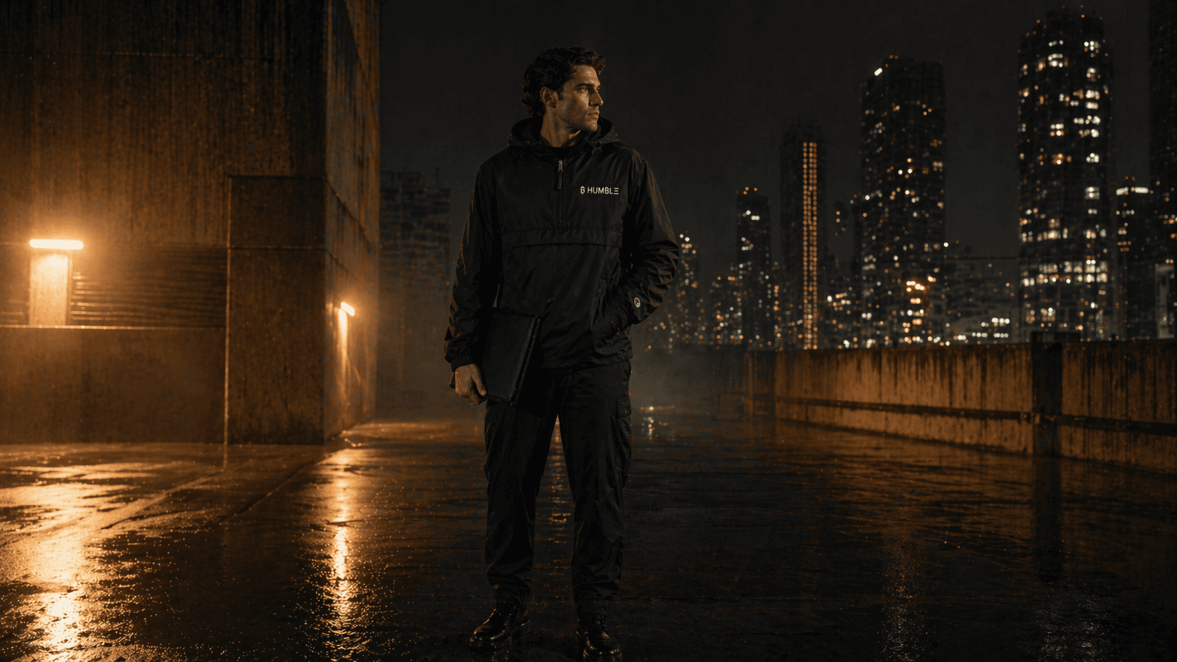 Editorial frame of a member standing in rain-soaked city light wearing matte-black ₿ HUM₿LΞ outerwear.